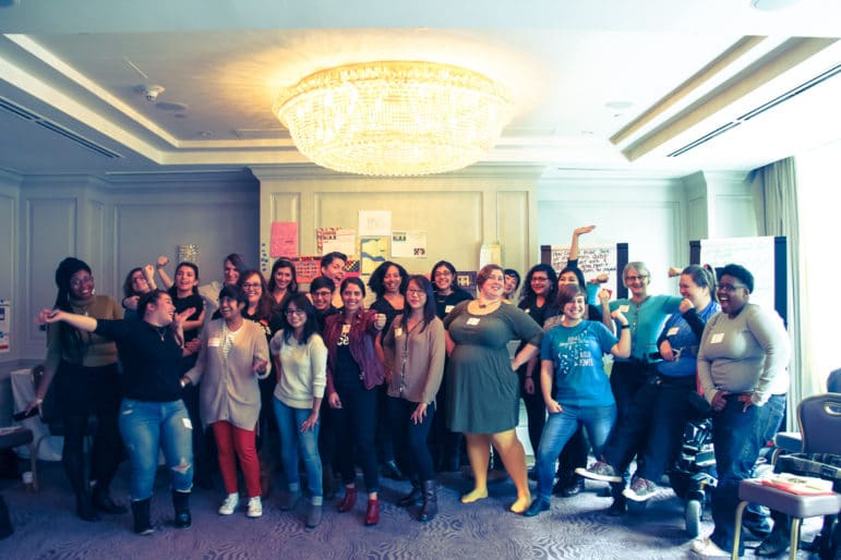 A large group of people pose together in a conference room, smiling and making strong poses under a chandelier.