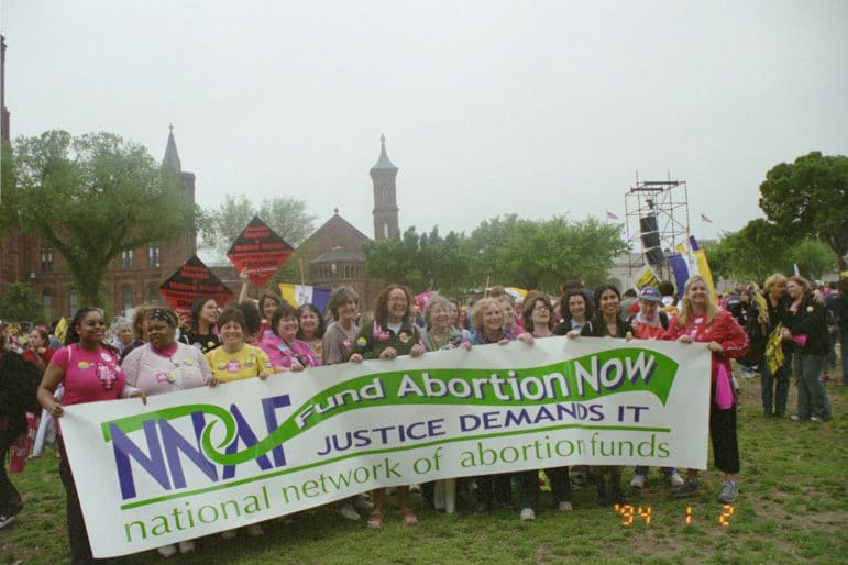 A large group of people stand together holding a banner that reads Fund Abortion Now, Justice Demands It, National Network of Abortion Funds.