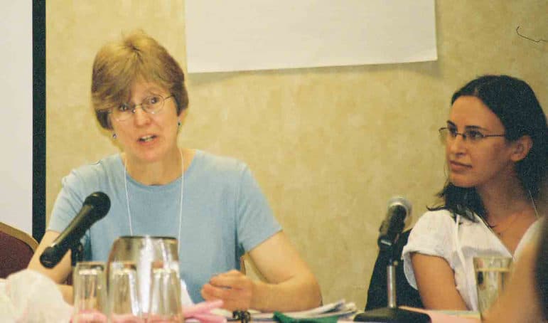 Two people sit at a table with microphones, one speaking and one listening.