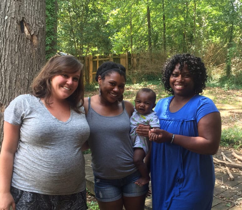 Three adults and a baby smile together outdoors on a sunny day.