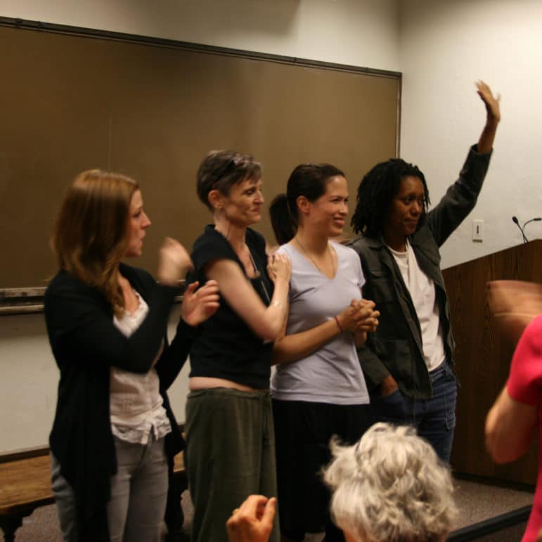 Four people stand together at the front of a room, smiling and gesturing as one waves to the audience.