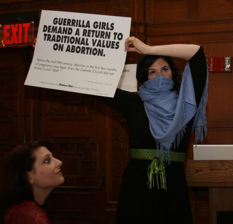 A person with a scarf covering their face holds a Guerrilla Girls sign calling for abortion justice, while another person looks on.