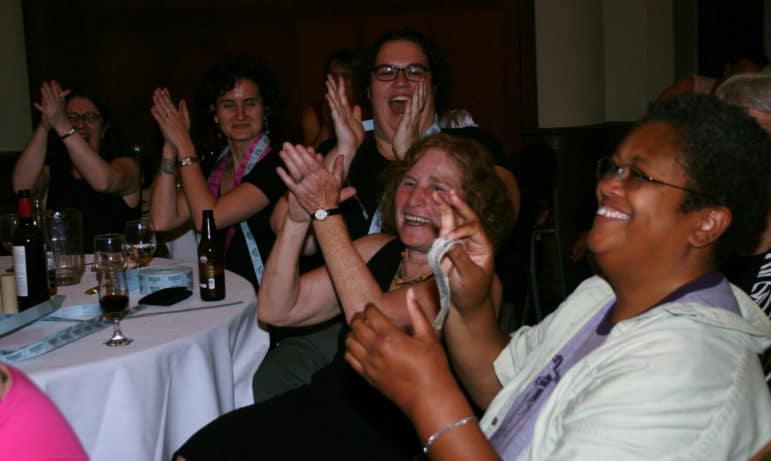 A group of people sit together at tables, clapping and laughing during an event.