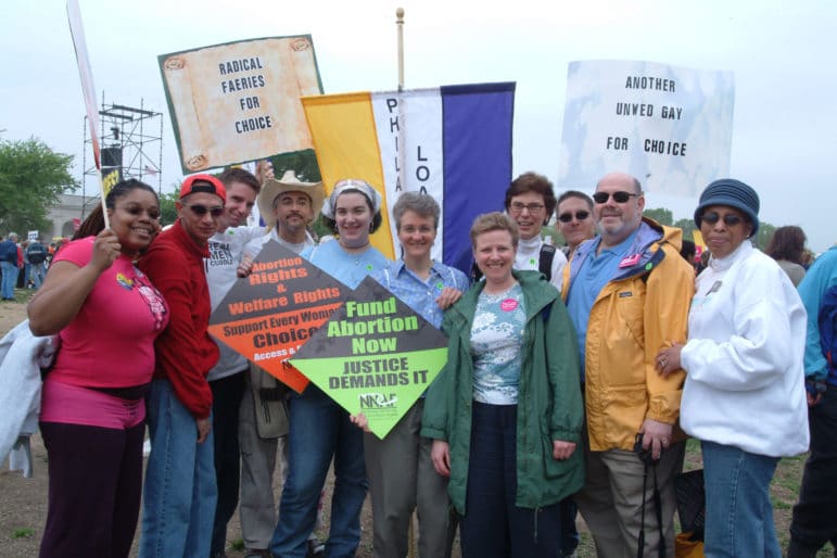 A group of people stand together outdoors holding colorful signs, including one that says Fund Abortion Now.