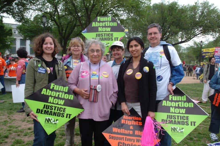 A small group of people hold bright signs that read Fund Abortion Now and Justice Demands It.